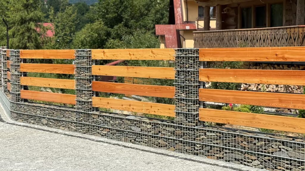 Ogrodzenie gabionowe Gablux z siatki spawanej o oczku 5x20 cm, wypełnione kamieniem, połączone z drewnianymi panelami w układzie naprzemiennym.